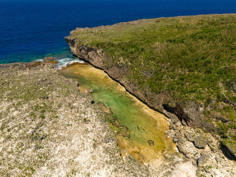 Aerial view of Chigit Beach featuring rugged limestone cliffs, turquoise shallow waters, and lush green vegetation in Northern Tinian, Tinian, Northern Mariana Islands.