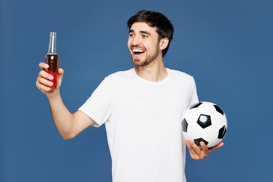 Young excited fun smiling man fan wear basic white t-shirt cheer up support football sport team hold in hand soccer ball watch tv live stream drink beer look aside isolated on plain blue background