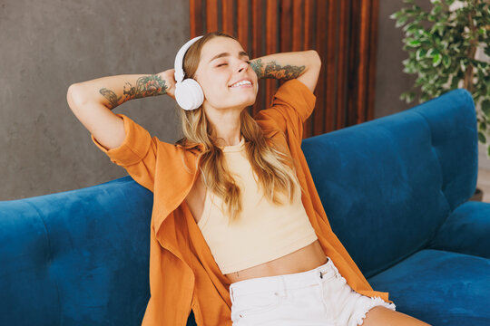 Young woman wears brown shirt headphones listen to music hold hands behind neck sit on blue sofa stay at home flat relax spend free spare time in living room indoors grey wall. People lounge concept.
