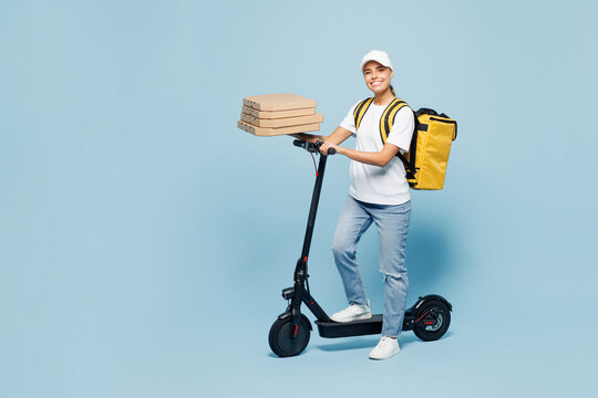Delivery woman wear white cap T-shirt uniform yellow thermal food bag backpack work as dealer courier ride e-scooter hold pizza in cardboard flatbox isolated on plain blue background. Service concept.