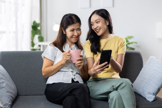 Asian mother and adult daughter relaxing on a sofa at home, happily viewing amusing content together on a smartphone.