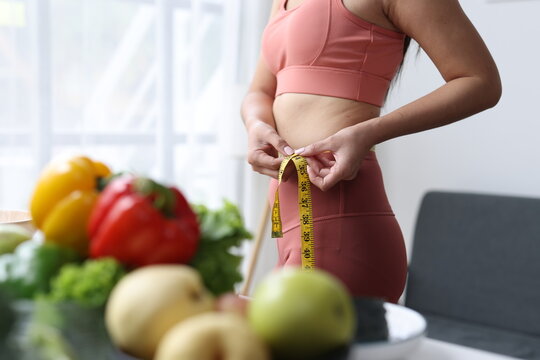 Woman standing in sportswear measuring her slim waist, focusing on body fitness and weight loss goals with fresh healthy vegetables and fruits on a table in the foreground.