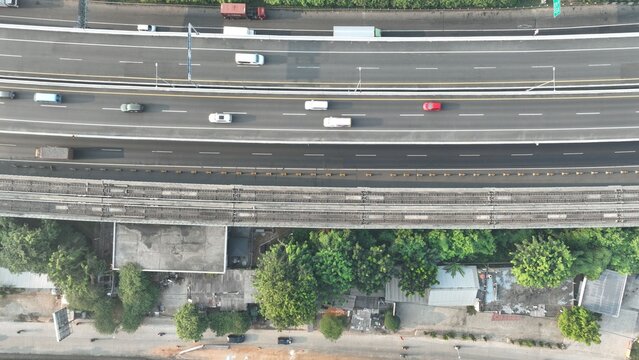 Aerial view of a multi-lane highway with cars and trucks, adjacent to a railway line and lush green trees in Bekasi Barat, West Java, Indonesia.