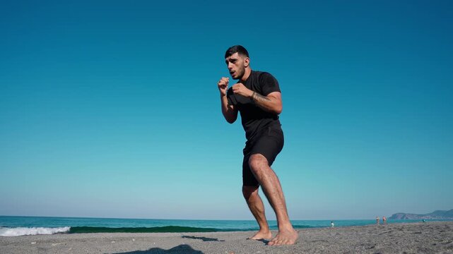 A man in black athletic wear practices boxing punches barefoot on a sandy beach by the sea under a clear blue sky.
