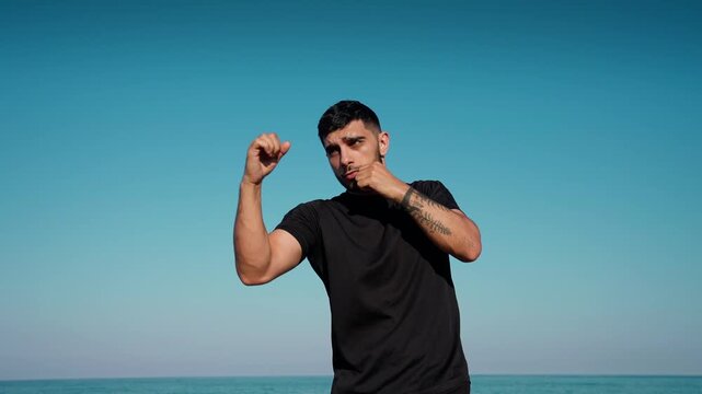 A young man wearing a black shirt training boxing shadowboxing moves under clear blue sky near the ocean.
