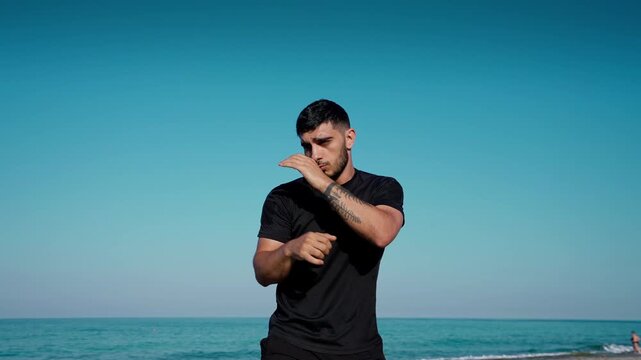A young man in a black shirt practices shadowboxing by the seaside with a clear blue sky and calm ocean behind him.
