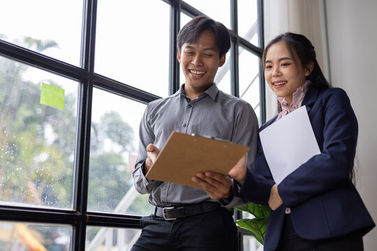 Professionals discussing business documents standing near window. Two smiling asian business people collaborating on a project, reviewing paperwork and documents in a office.