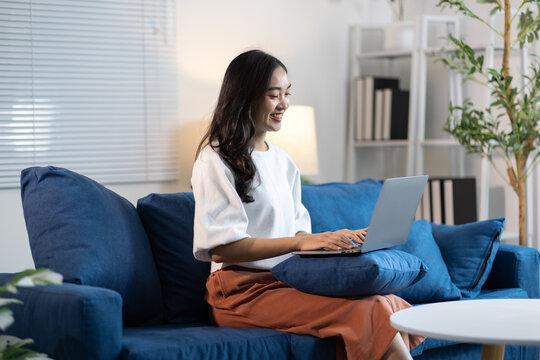 Happy Asian woman smiling, confidently working on laptop from a comfortable home sofa.