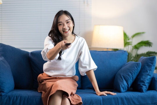 Happy young Asian woman watching TV at home. Young Asian woman smiling, holding a remote control, sitting on a blue couch in her living room, enjoying entertainment.