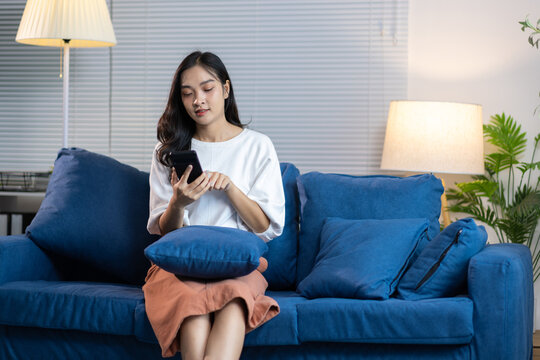 Smiling woman sitting on a sofa, holding a mobile phone, and enjoying leisure time in her cozy living room.