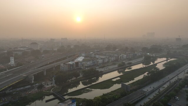 Aerial view of the urban landscape featuring a river, elevated highway, and buildings under a hazy sunset sky in Bekasi Barat, West Java, Indonesia.