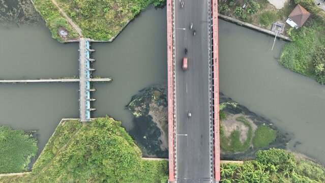 Aerial view of a bridge crossing a river with a small weir structure and lush green vegetation in Bekasi Barat, West Java, Indonesia.