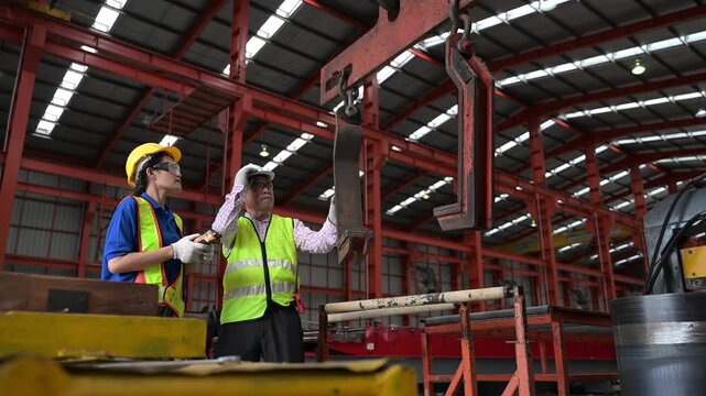 Two professional industrial engineers wearing safety helmets and uniforms monitoring the overhead crane operation and discussing safety protocols near sheet metal rolls in a factory plant.
