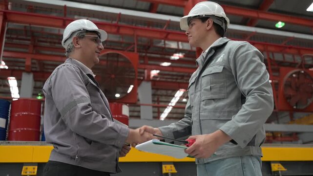 Two professional industrial engineers wearing safety helmets and uniforms shaking hands and holding a clipboard during a strict safety audit in a manufacturing factory warehouse.