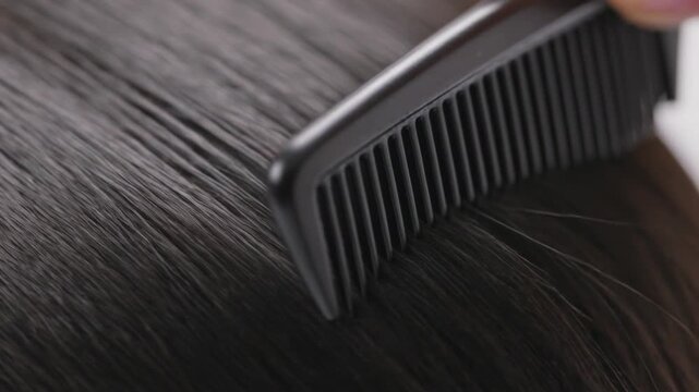 Combing hand with black plastic comb moving from right across scalp at salon, smoothing hair part