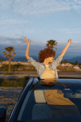 Obraz premium A woman enjoys a carefree moment from a car roof, arms raised, in a sunny outdoor setting. Lifestyle concept capturing joy, freedom, and casual travel.