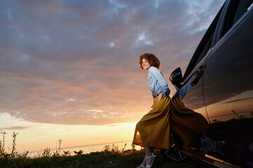 Obraz premium Woman with curly hair in a denim shirt and mustard skirt stands by a car at sunset, capturing a lifestyle moment with outdoor fashion, relaxed vibe and travel spirit in warm light.