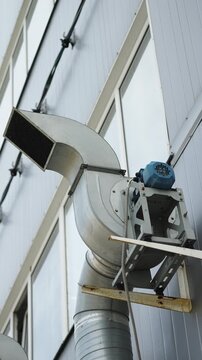 Exterior industrial ventilation system with galvanized ductwork and centrifugal exhaust fan mounted on a building facade, featuring curved elbow, grille outlet, and support brackets.