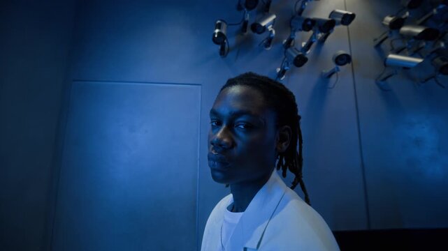 Tilt up portrait of young African American man standing under multiple CCTV surveillance cameras in security room in blue light. For concepts on cybersecurity, digital privacy, and AI monitoring