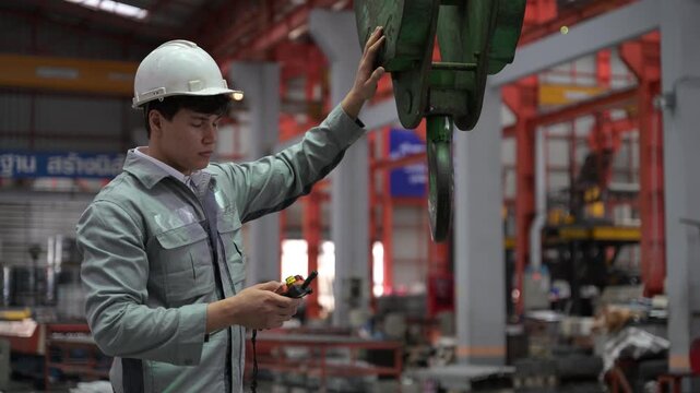 An industrial worker wearing protective gear guides and secures a green crane clamp hook mechanism over steel materials in a factory.