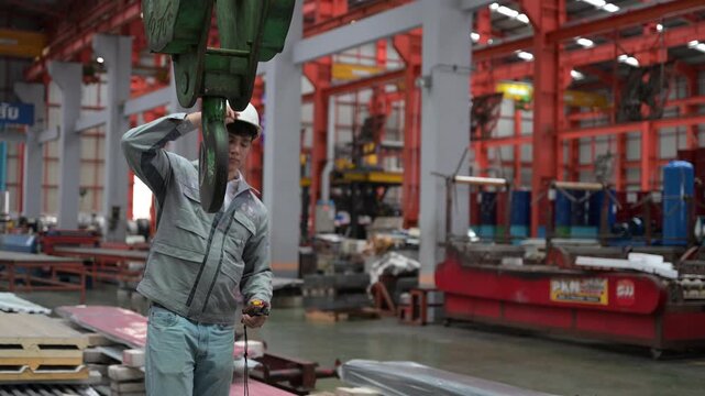 An industrial worker wearing protective gear guides and secures a green crane clamp hook mechanism over steel materials in a factory.