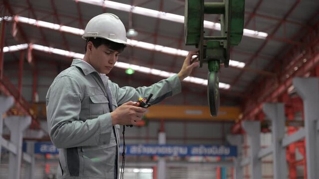 An industrial worker wearing protective gear guides and secures a green crane clamp hook mechanism over steel materials in a factory.
