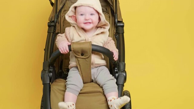 Happy child sitting in stroller on yellow background. Small baby in warm clothing resting in baby carriage. Infant leisure outdoors or walk, parenting concept, childhood growth.