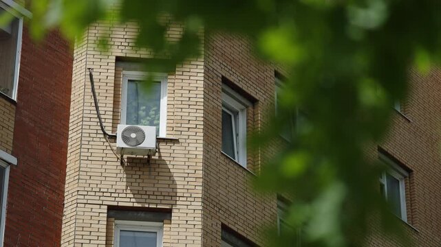 Corner of a residential apartment building with beige brick facade, white windows, and an exterior air conditioner unit, framed by blurred green leaves on a bright summer day