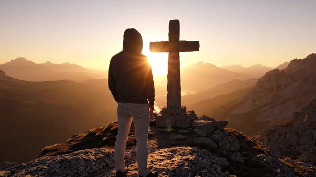 Man standing before stone cross on mountain peak at golden sunset