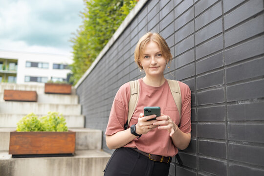 A young happy blonde woman in a pink t-shirt and jeans stands on a city street near a stone wall, looking at her smartphone and smiling on a sunny day.