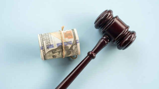 Wooden judge gavel laying next to a roll of US one hundred dollar bills on solid plain blue background for legal expense and corruption theme