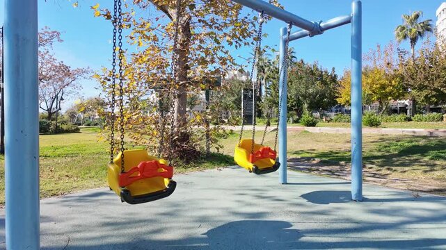 Two empty yellow baby swing is swinging on chains in sunny Antalya park with green trees.