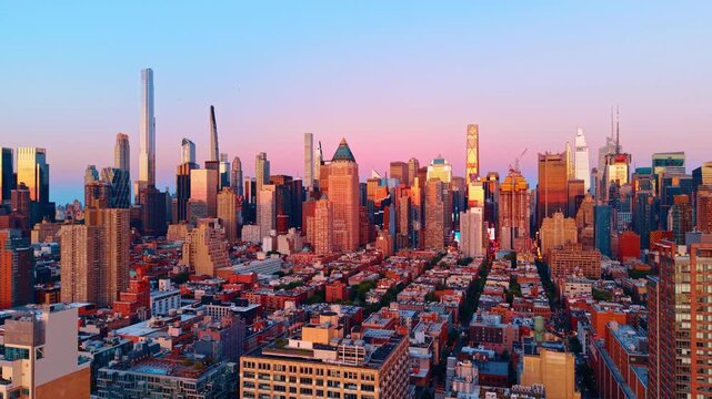 Midtown Manhattan skyline at sunset from the west side, New York. Panoramic elevated view of mixed residential and commercial towers bathed in pink and orange light under a blue-to-rose gradient sky.