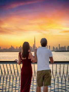 A romantic tourist couple stands at Creek Harbour and looks at the distant skyline of Dubai, UAE, during a beautiful sunset