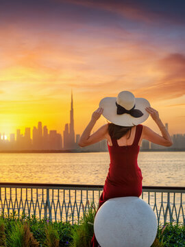 A beautiful woman in a dress and with hat looks at the distant skyline of Dubai, UAE, during sunset time