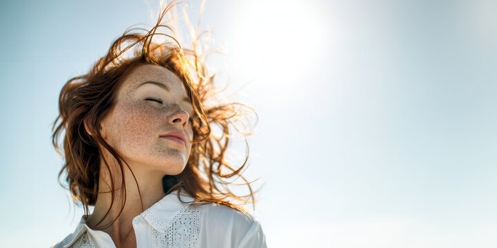 Sensual young woman with long, vibrant, wavy curly red hair and natural freckles, closed eyes peacefully, standing, relaxing, enjoying under the bright warm glow of the summer sun outdoors,  copyspace