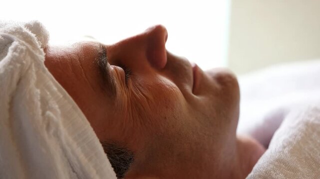 Mature man relaxing with a facial treatment at a luxury spa resort, lying comfortably on a massage table with a white towel wrapped around head