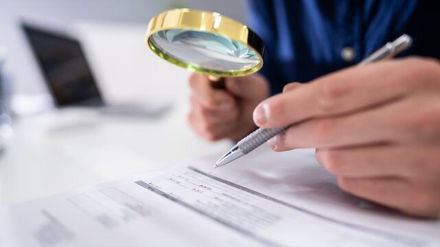 Businessman examining documents with magnifying glass, office desk with laptop, close-up hands holding pen and loupe