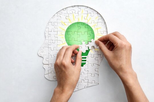 Vista superior de manos colocando piezas blancas de rompecabezas sobre una silueta de cabeza humana con una bombilla verde brillante. Concepto de psicolog&iacute;a, ideas creativas y salud mental.