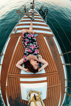 Overhead fisheye view of brunette woman in black floral dress lying on sailboat bow deck, arms up, smiling, surrounded by teak wood deck and calm teal sea water