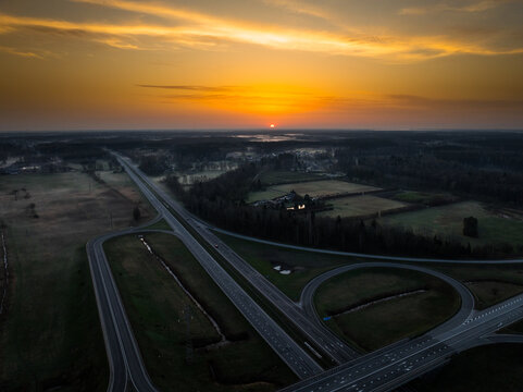 Aerial view of the A5 and A8 highway interchange at sunset, featuring sweeping roads, surrounding fields, and a vibrant orange sky in Olaine parish, Olaine Municipality, Latvia.