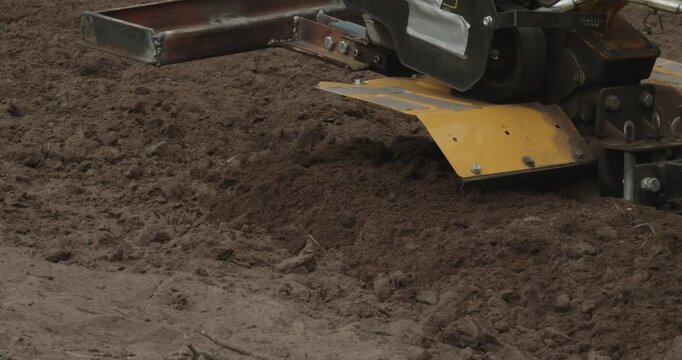 Farmer using a rototiller cultivator to plow the ground and prepare the soil for planting new crops in the garden, showcasing modern agricultural machinery at work on a farm field