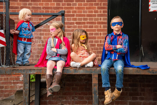 Four children in superhero costumes sitting on a brick platform.
