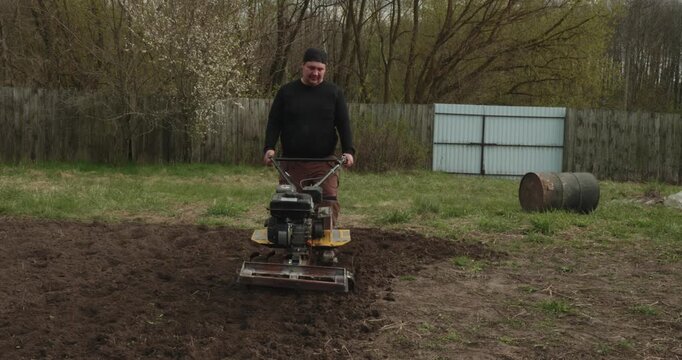 Male farmer using a motor cultivator to till the ground in his backyard, preparing the soil for planting seeds during the spring agricultural season with specialized farming equipment
