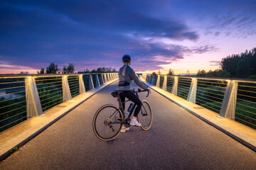 Naklejka premium Female cyclist with road bike on modern illuminated bridge at sunset in Slovenia. Woman riding bicycle in helmet on asphalt path, active sport lifestyle, beautiful twilight sky landscape. Girl on bike