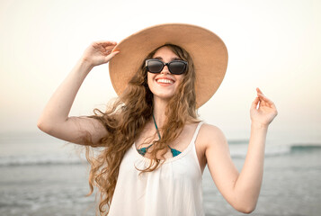 Smiling young woman with windblown hair and sunglasses holding sun hat on beach, happiness, joy, positive emotions, carefree summer lifestyle and wellbeing concept © Beatriz