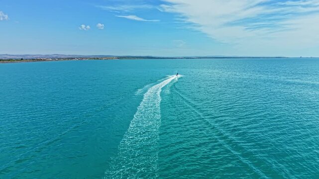 A boat travels across calm water during daylight, leaving a trail of waves behind. The sky is blue with some clouds and land is visible in the distance.