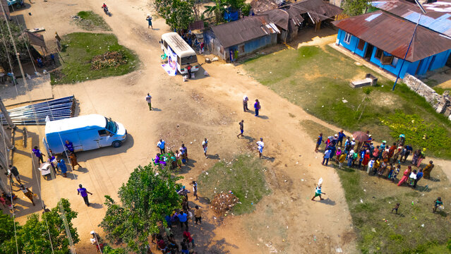 Aerial view of a community gathering with a white van and a small bus parked on a dirt road, surrounded by houses with metal roofs and groups of people in Abua, Rivers, Nigeria.