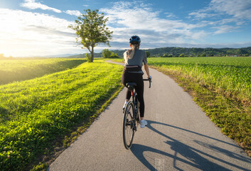 Fototapeta premium Female cyclist with road bike riding on asphalt path at sunset. Summer in Slovenia. Rear view of woman cycling in helmet near green fields and tree, active sport lifestyle, evening sun sky landscape