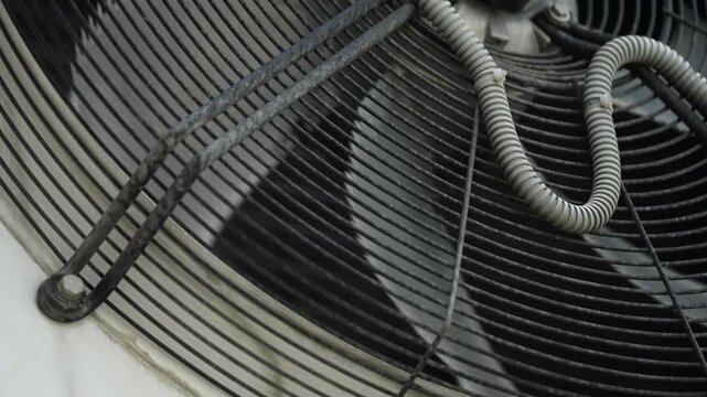 Close-up view of an industrial HVAC condenser fan with spinning blades behind a protective metal grille, coiled electrical conduit across the guard, highlighting cooling airflow